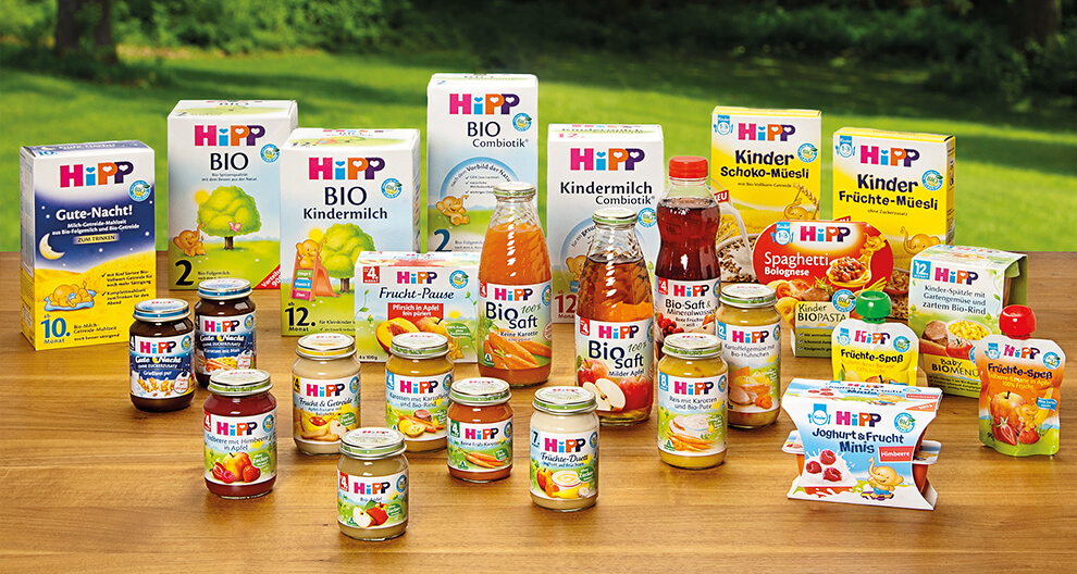 Various Hipp baby food products displayed on a wooden table with a green grassy background.