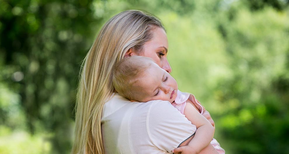 A woman with long blonde hair holds a baby against a blurred green background.