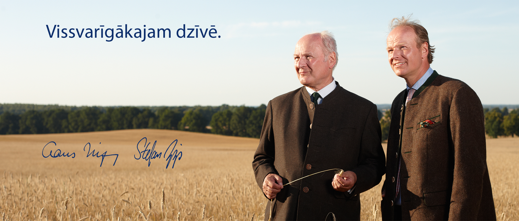 Divi vīri uzvalkos stāv kviešu laukā, kura ainaviskais fons ir koki un skaidras debesis.