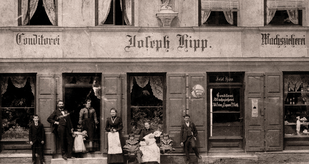 Grupa cilvēku, tostarp pieaugušie un bērni, stāv un sēž pie veikala ar uzrakstu "Johans Hipps".