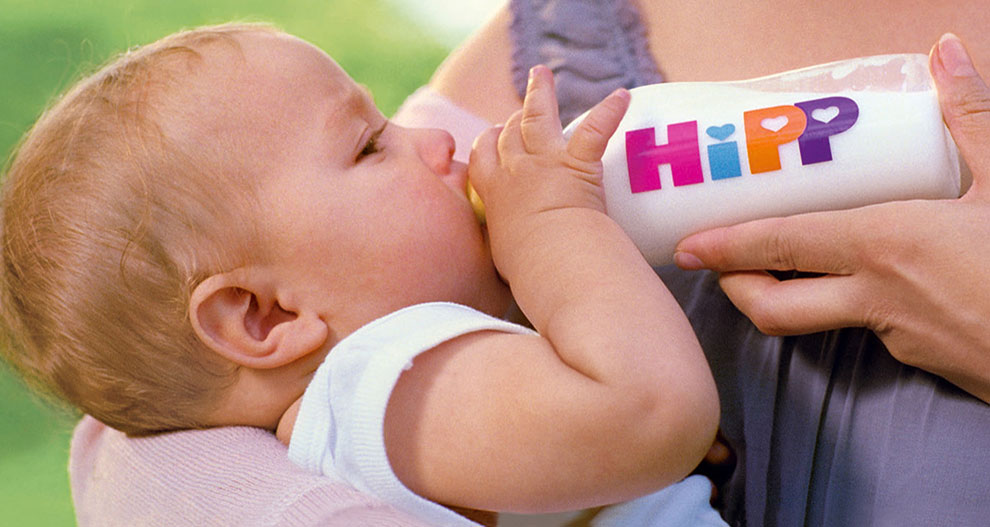 Bērns dzer no pudeles ar uzrakstu "HiPP".