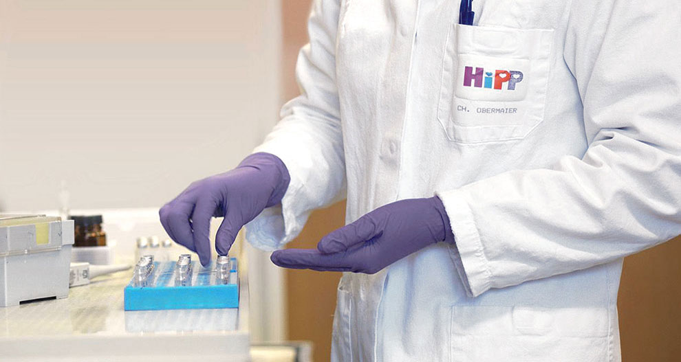 Persona baltā laboratorijas halātā ar logotipu "HiPP" un nosaukumu "CH. OBERMAIER", iespējams, laboratorijas apstākļos, tur zilu paplāti ar nelieliem priekšmetiem.