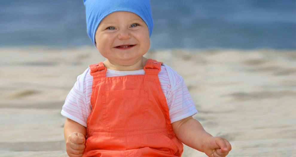 Bērns sēž smilšainā pludmalē, zilā cepurē un oranžā kombinezonā.