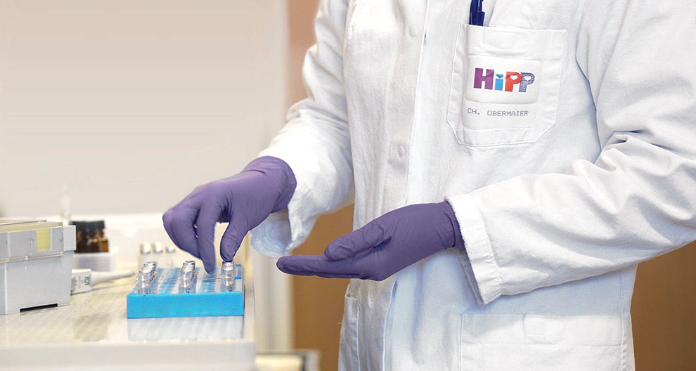 Persona baltā laboratorijas halātā ar logotipu "HiPP" un nosaukumu "CH. OBERMAIER" tur zilu paplāti ar mazām flakoniņām.