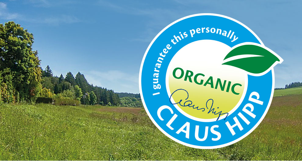 Ainava ar zaļu zāli un kokiem ir pārklāta ar apaļu grafiku, kurā ir uzraksts "Es personīgi garantēju" un "CLAUS HIPP" ar lapas simbolu un vārdu "ORGANIC" centrā.