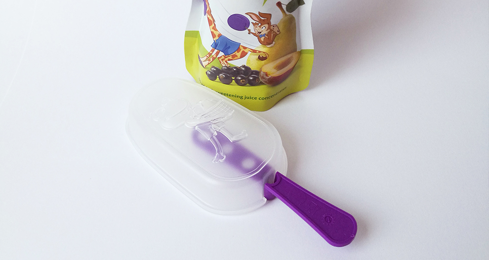 Plastmasas saldējuma veidne ar violetu rokturi un sulas maisiņš ar augļu ilustrāciju.